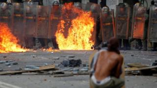 Hombre cubriéndose con un escudo casero contra funcionarios de la Guardia Nacional Bolivariana.